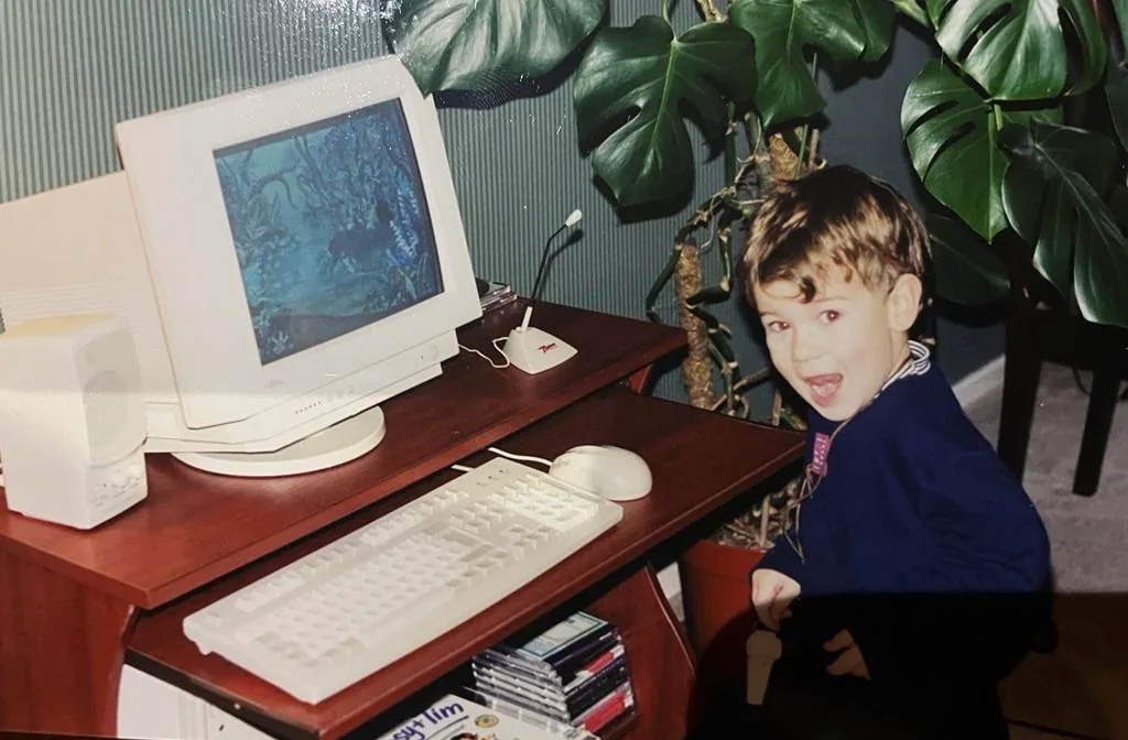 Ben in 1997, aged three, grinning over his shoulder at the camera beside his first computer: a beige Fujitsu monitor showing an underwater game, a beige keyboard and mouse, a desk speaker and microphone, a stack of CD-ROMs under the desk and large monstera plants behind