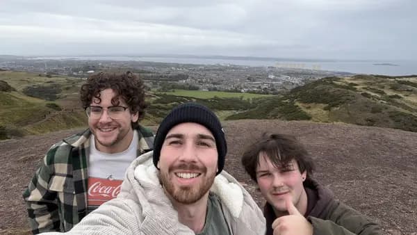 Three young men smiling in a selfie on the top of Arthur's Seat on an overcast day, the one in the middle in a dark beanie, with Edinburgh and the Firth of Forth spread out behind them.