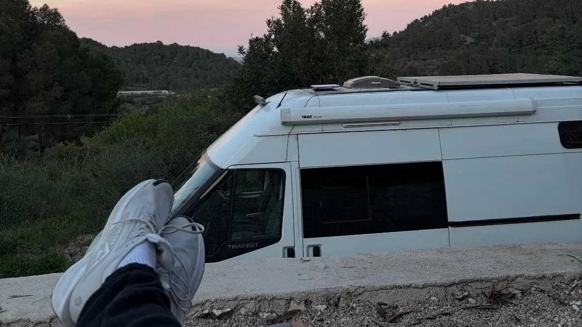 A white Ford Transit campervan with a solar panel and a Starlink satellite dish on its roof parked below pine-covered hills in south-east Spain at sunset, with a pair of white trainers resting on a low wall in the foreground under a pink sky.
