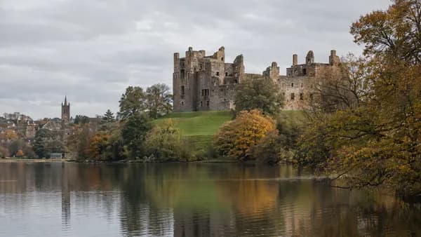 A ruined royal palace of pale stone on a grassy rise above a calm loch on a still autumn day, with trees turning colour along the shore.