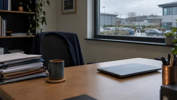 A small office unit on a business park with a desk, a laptop and a stack of paperwork by the window on a grey day.