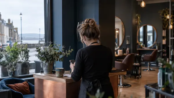A salon owner seen from behind tests a booking form on a phone at a reception desk in Musselburgh.