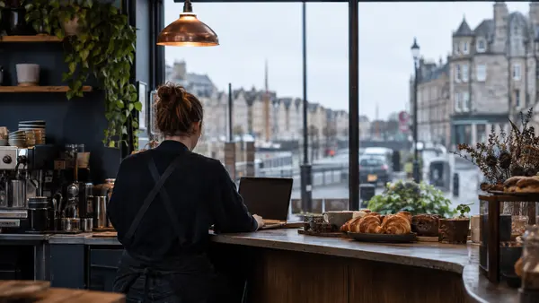 A cafe owner viewed from behind uses a laptop beside a counter in Leith.