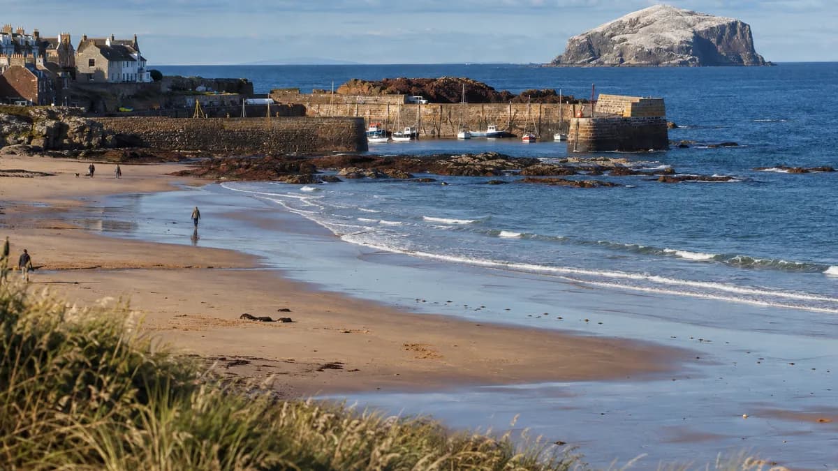 A sandy beach and a small harbour on the East Lothian coast on a sunny day, with a steep rocky island far out to sea.