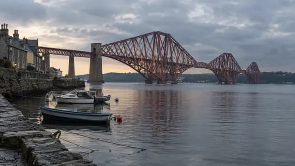 The red steel spans of the Forth Bridge seen from the waterfront at South Queensferry on a calm grey evening, with small boats moored in the foreground.