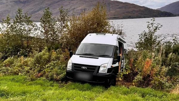 A white campervan nose-down in bushes at the edge of Loch Lomond, hills on the far shore under a grey sky.