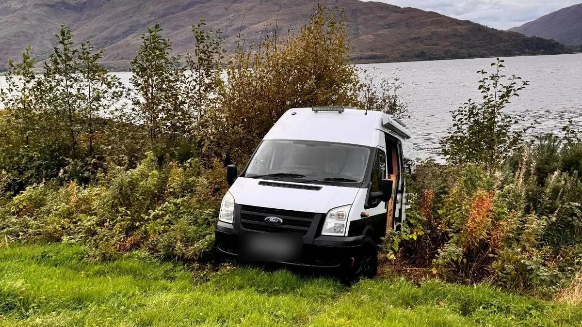 A white campervan nose-down in bushes at the edge of Loch Lomond, hills on the far shore under a grey sky.