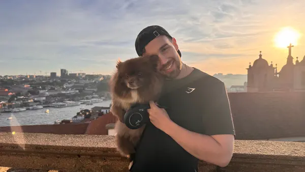 A smiling man in a black cap and black t-shirt holds a fluffy brown Pomeranian on a stone terrace in Porto at sunset, with the Douro river and the city's rooftops behind him and a church tower silhouetted against the sun.